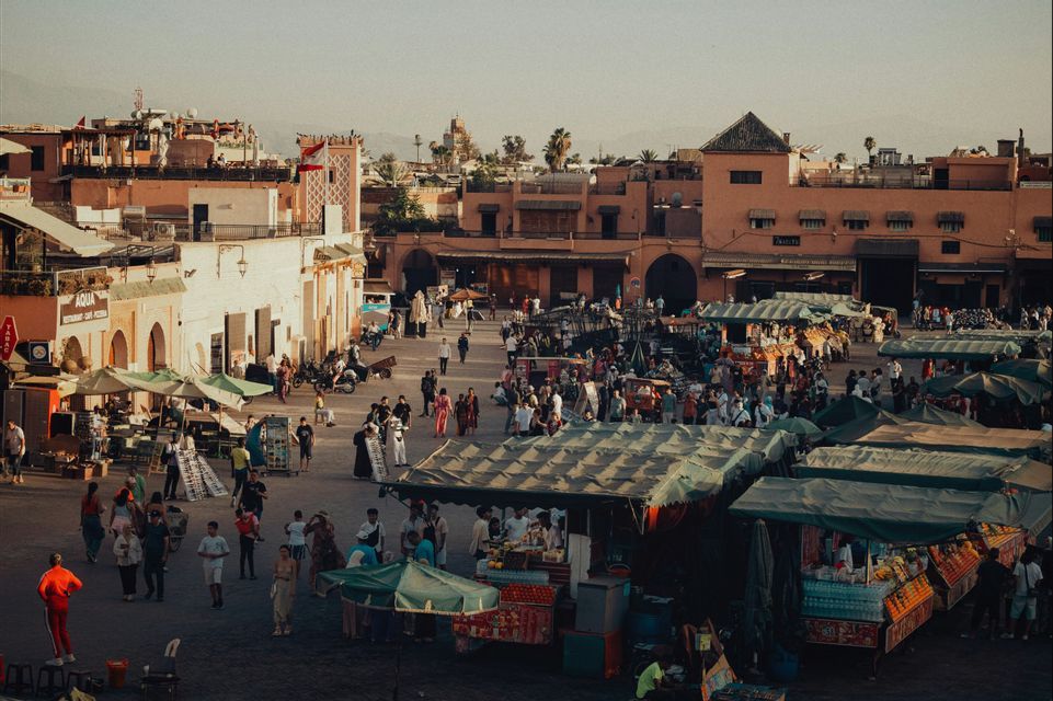 Vue plongeante d'une place de ville animée, remplie de gens et d'étals de marché avec des auvents verts au coucher du soleil.