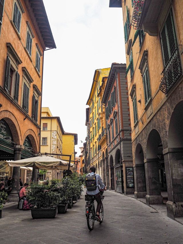Ein Mann mit Rucksack fährt mit dem Fahrrad eine enge Straße entlang, die von hohen, farbenfrohen Gebäuden mit Bogengängen gesäumt ist.