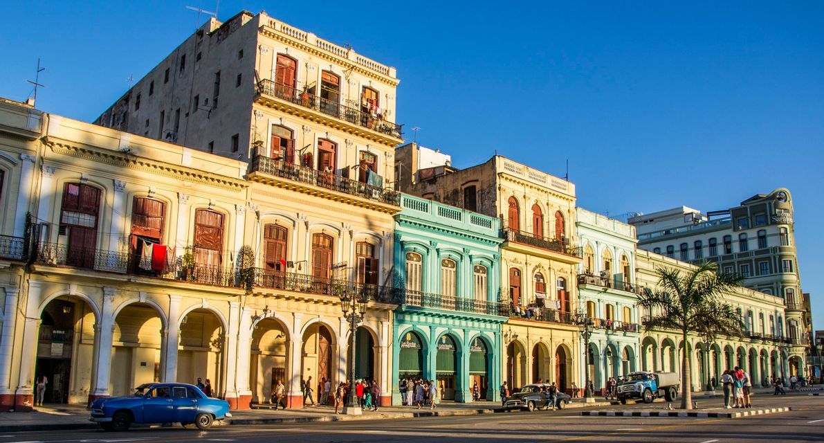 Des bâtiments colorés de style colonial bordent une rue de la ville où des gens se promènent et des voitures anciennes circulent sous un ciel bleu clair.