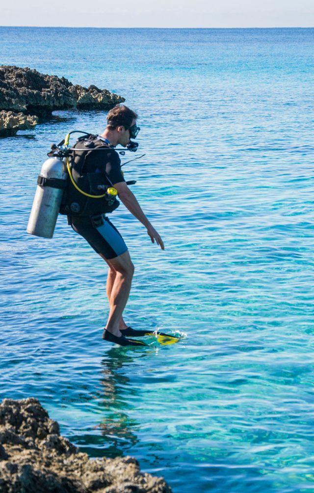 Un hombre con equipo completo de buceo se prepara para entrar en el mar azul claro desde una costa rocosa.