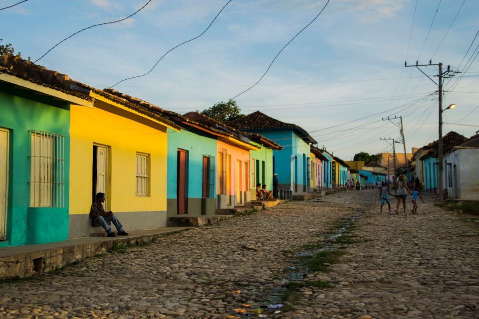 Una calle empedrada bordeada de casas coloridas en verde, amarillo y azul, con gente relajándose al aire libre durante el atardecer.