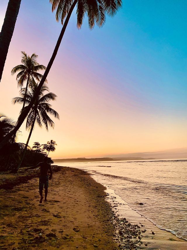 Eine Person spaziert an einem sandigen Strand unter Palmen, mit einem farbverlaufenden Sonnenuntergangshimmel über dem Meer.