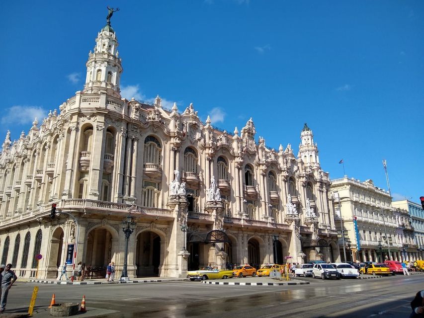 Un edificio classico, ornato e di colore chiaro, con torri, si erge all'angolo di una strada cittadina. Davanti, sotto un cielo azzurro, sono parcheggiati taxi gialli d'epoca e altre auto.