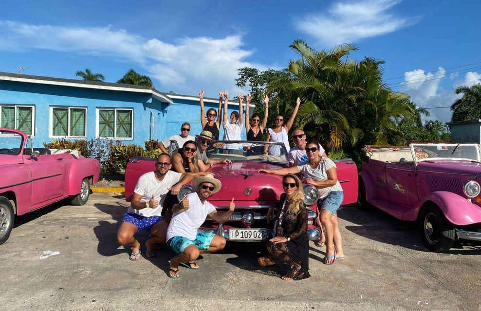 Un gruppo WeRoad in viaggio sorride e posa per una foto intorno a una decappottabile rosa d'epoca, davanti a un edificio blu.