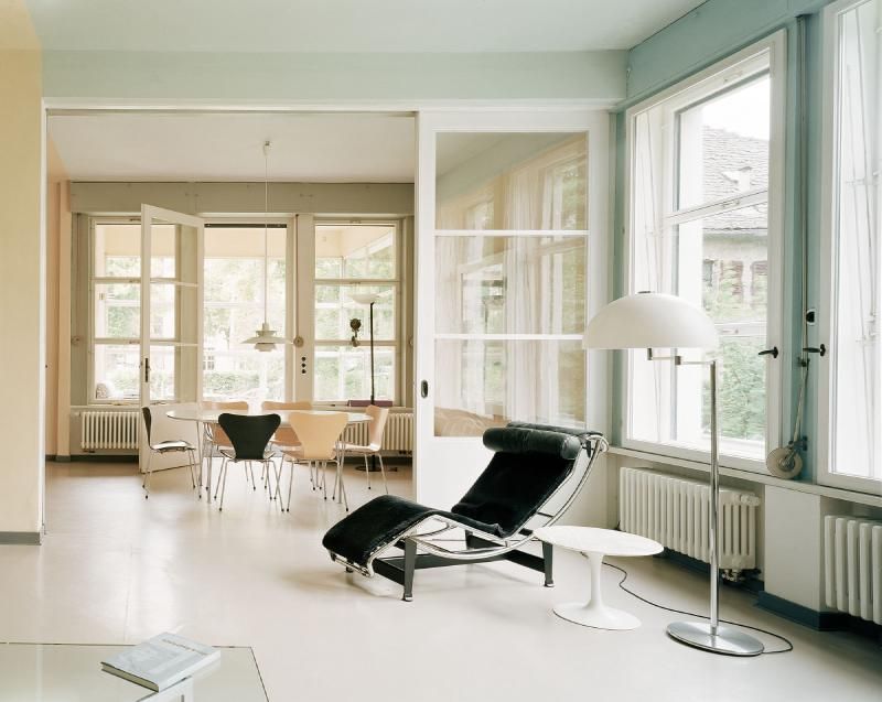 Una camera moderna con pavimento bianco, una chaise longue nera e una zona pranzo situata dietro un'ampia apertura.