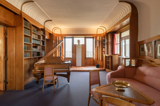 Una camera Art Nouveau con ampie boiserie in legno, un pianoforte a coda, librerie a muro e un divano rosa su un tappeto blu.