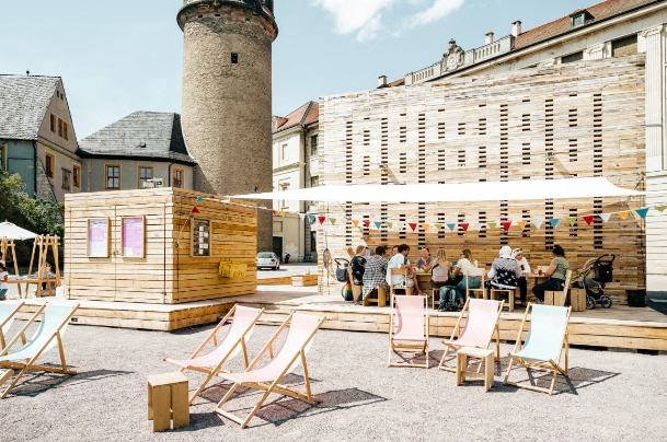 Un gruppo WeRoad a un tavolo in un cortile soleggiato, con strutture in legno temporanee e sedie a sdraio colorate.