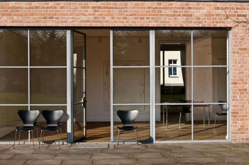 Un edificio in mattoni con una parete di finestre a tutta altezza, una porta aperta e tre moderne sedie nere sul patio.