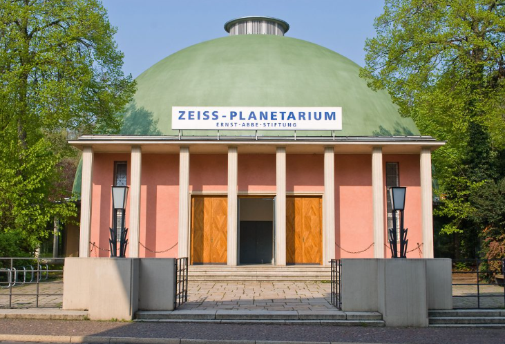 L'ingresso principale dell'edificio Zeiss-Planetarium, con la sua grande cupola verde, il portico a colonne e le porte in legno sotto un cielo sereno.