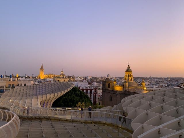 Menschen gehen auf einem modernen Hochweg mit Blick auf eine Stadt-Skyline mit beleuchteten Türmen bei Sonnenuntergang.