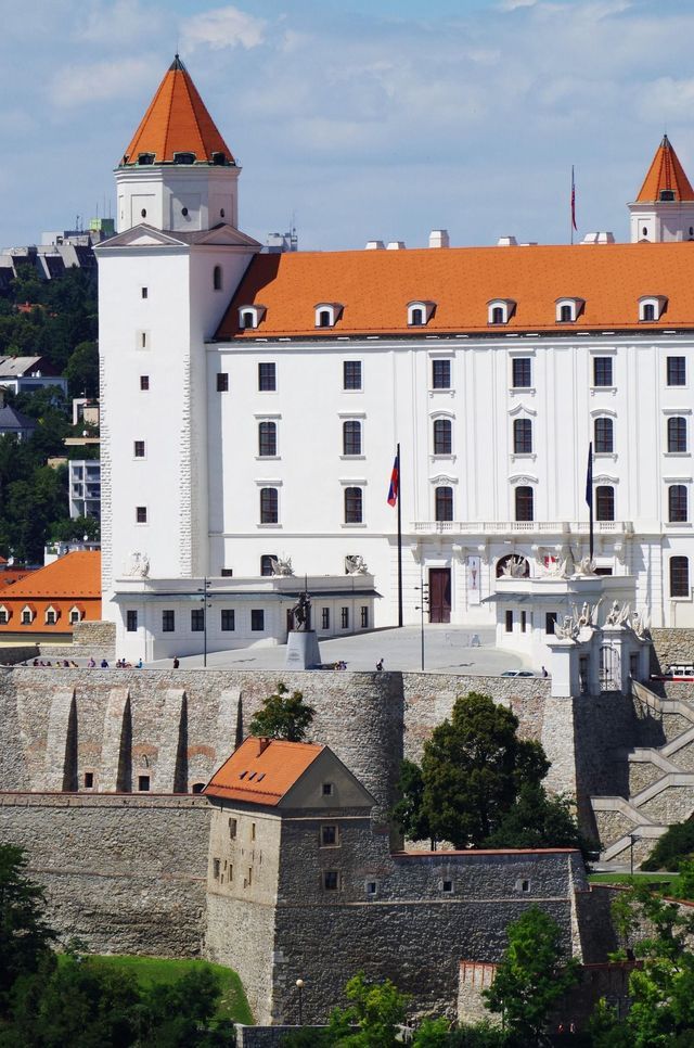 Un gran castillo blanco con techos de tejas naranjas y torres en las esquinas se asienta sobre una colina de piedra fortificada bajo un cielo parcialmente nublado.