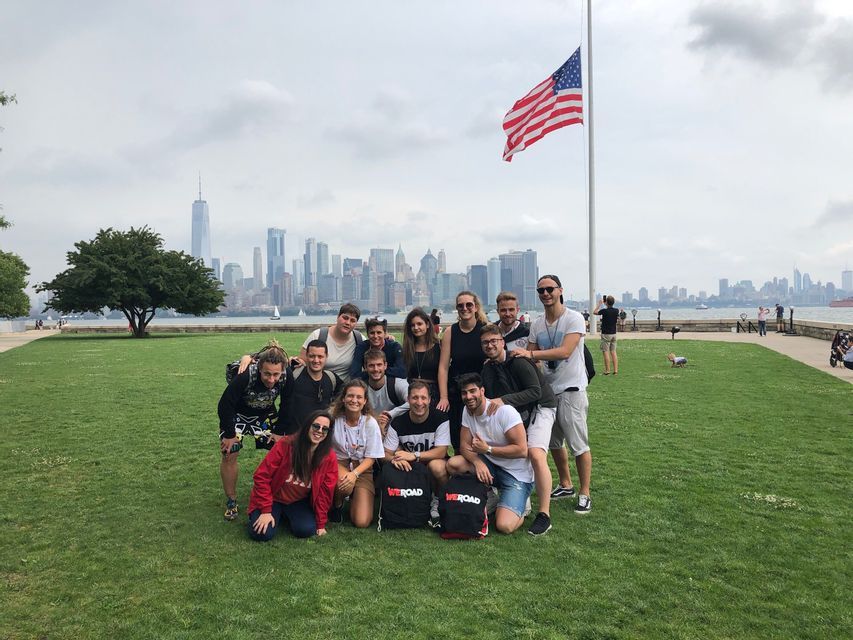 Un grupo de WeRoad posa para una foto en un césped con una bandera estadounidense y el horizonte de una ciudad de fondo.