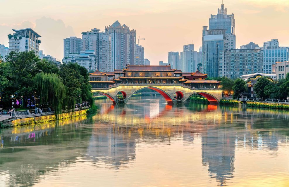 An illuminated traditional bridge with red arches spans a river, reflecting on the water with a modern city skyline in the background.