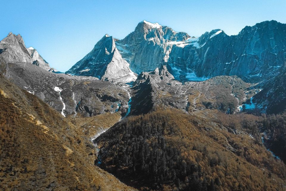 Jagged, snow-capped mountains tower over a valley with autumn-colored forests under a clear blue sky.