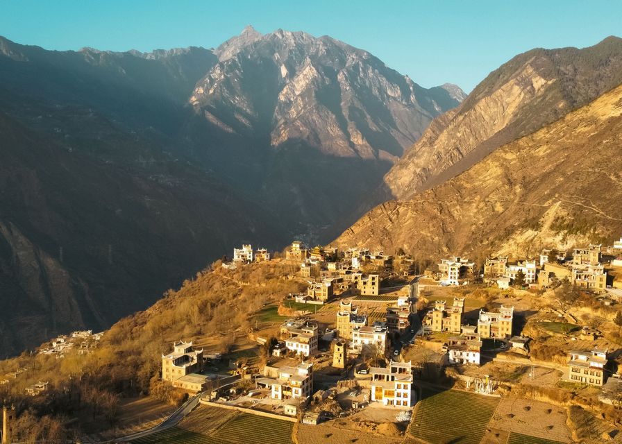 A village of traditional, multi-story buildings clings to a sunlit hillside at the base of a rugged mountain range.
