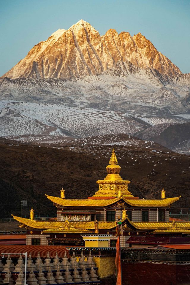 A golden temple sits in the foreground with a massive, snow-dusted mountain range illuminated by sunlight in the background.