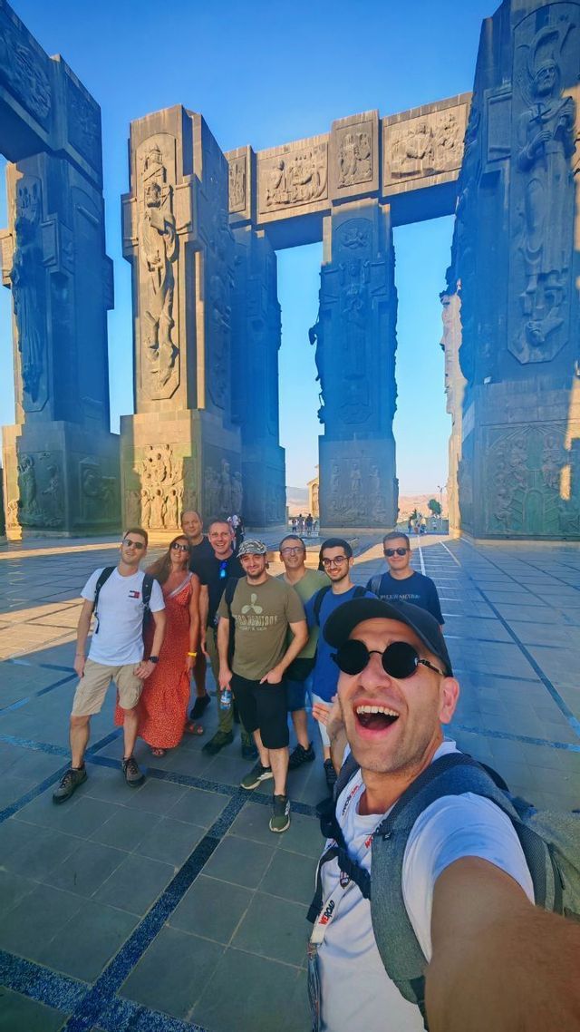 Un gruppo WeRoad scatta un selfie sorridente davanti a un grande monumento in pietra scolpita sotto un cielo sereno.