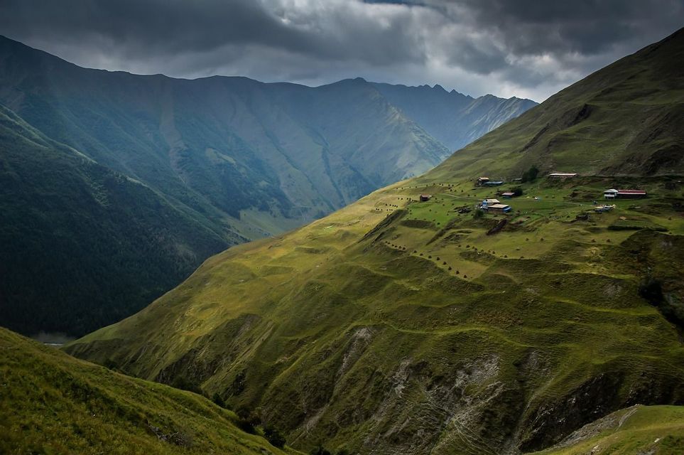 Un piccolo villaggio su un versante montuoso verde e soleggiato si affaccia su una vasta valle sotto un cielo nuvoloso.