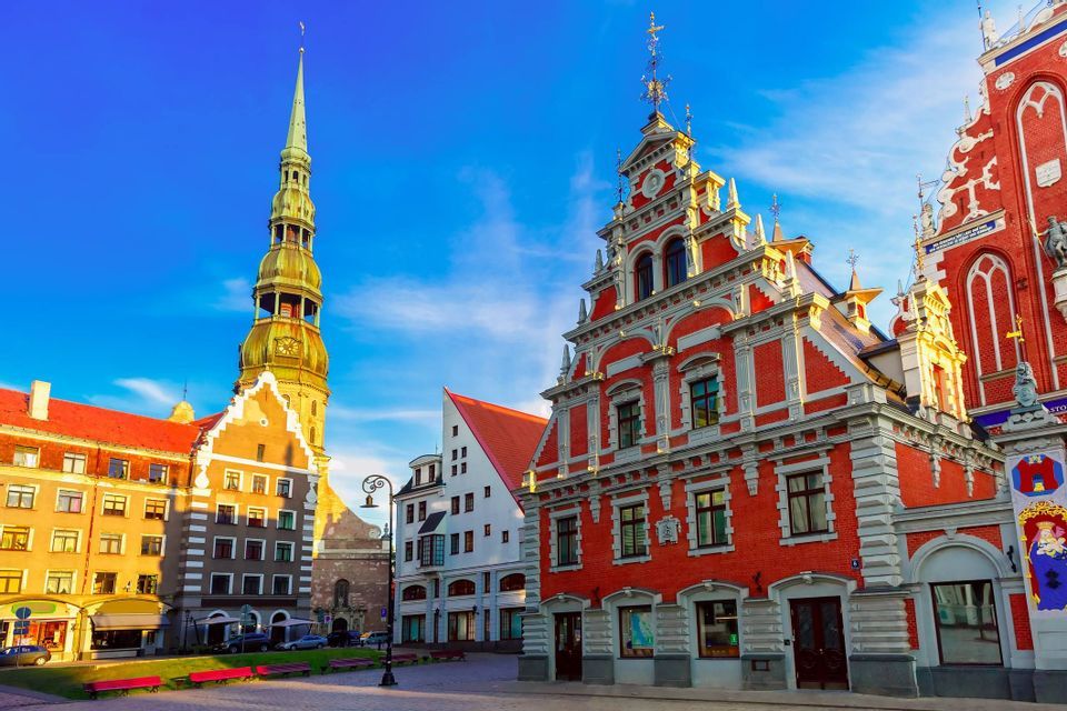 Ornate red brick buildings and a church spire stand in a city square under a clear blue sky.