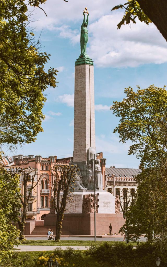 Un grand obélisque en pierre surmonté d'une statue, vu depuis un parc avec des arbres verts encadrant le monument sur fond de ciel bleu.