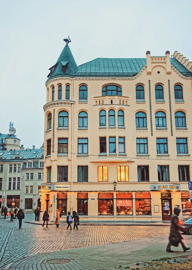 A yellow corner building with a black cat statue on its roof turret, located on a cobblestone street with people walking by.