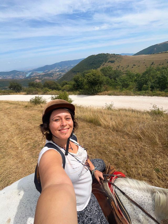 Una donna sorridente con un casco si scatta un selfie mentre cavalca un cavallo bianco su una collina, con una valle e un lago sullo sfondo.