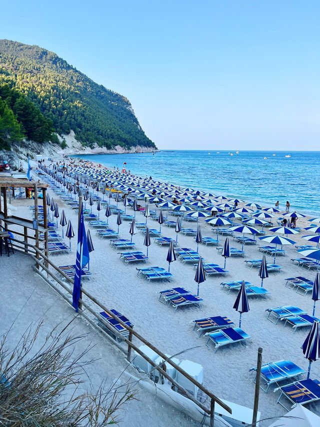 Una veduta dall'alto di una lunga spiaggia con file di ombrelloni e lettini blu e bianchi in riva al mare.