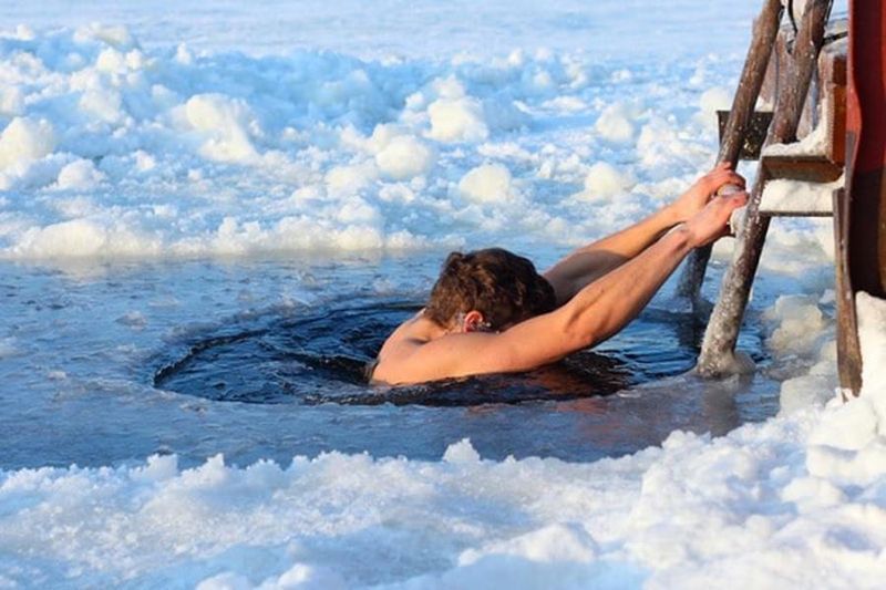 A person holds onto a wooden ladder while dipping into a hole cut into a frozen, snow-covered body of water.