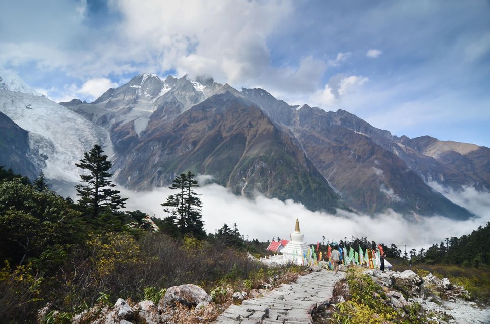 A stone path leads to a white stupa and prayer flags, with snow-capped mountains and a glacier in the background.