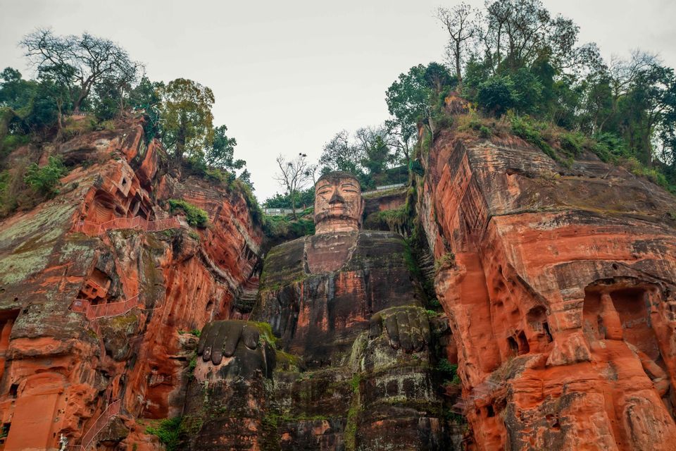 A giant Buddha statue carved into a red sandstone cliff, nestled between two rock faces with trees on top.