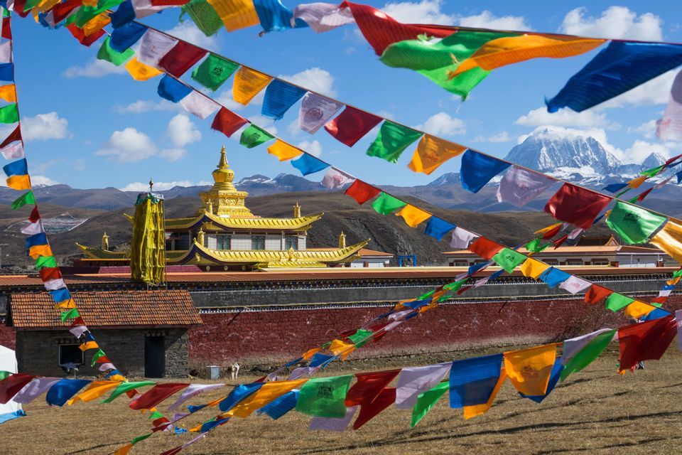 Colorful prayer flags blow in the wind, framing a view of a golden-roofed monastery and distant snow-capped mountains.