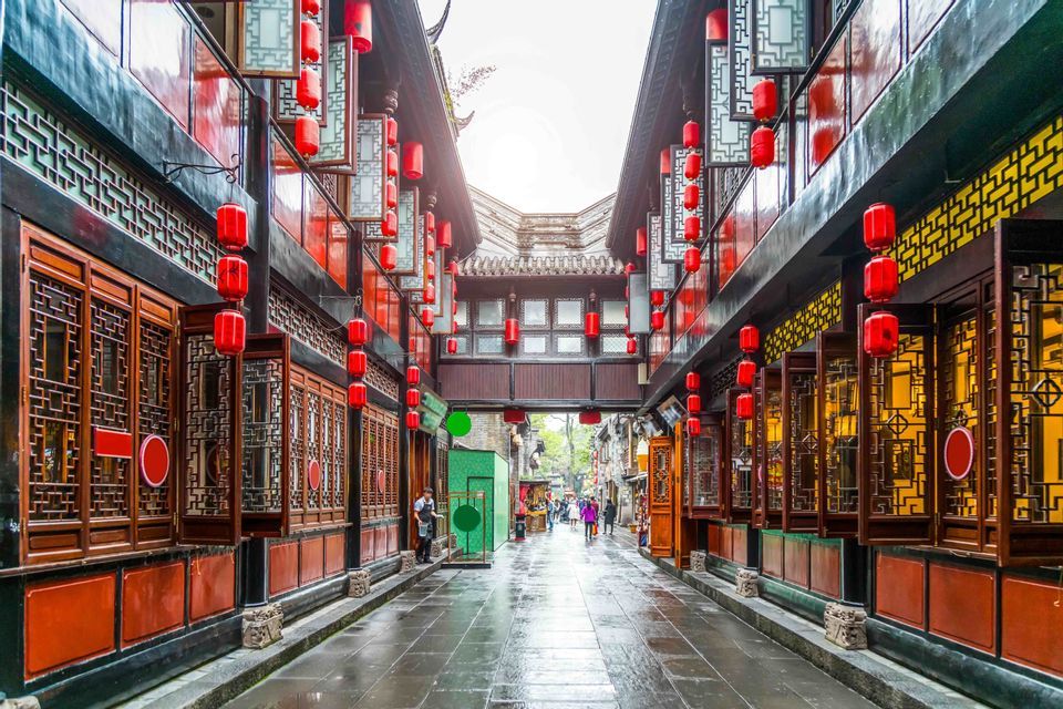 A narrow street with traditional architecture is lined on both sides with hanging red lanterns, reflecting on the wet pavement.