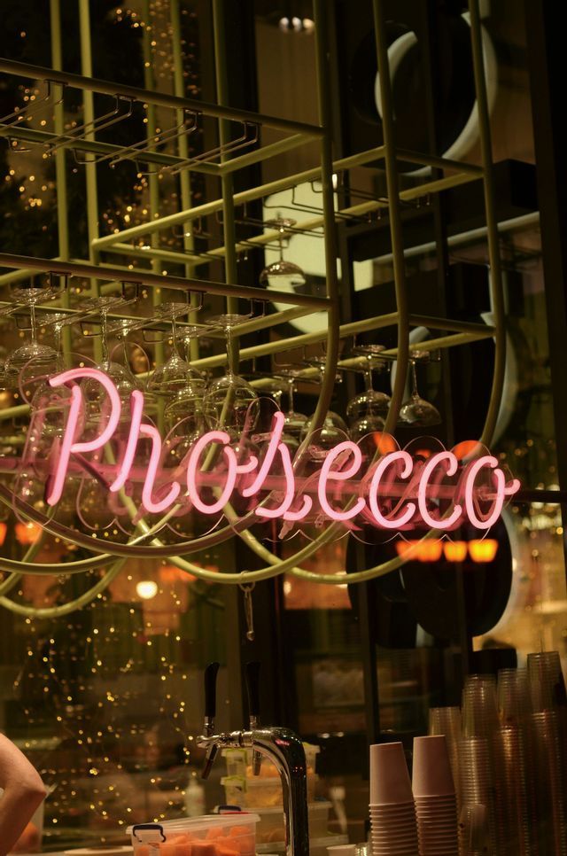 Un'insegna al neon rosa illuminata con la scritta 'Prosecco' all'interno di un bar, con calici da vino appesi a testa in giù a un portabicchieri.