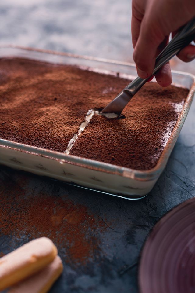Una mano taglia una fetta di tiramisù in una coppa di vetro, rivelando il ripieno cremoso sotto il cacao in polvere.