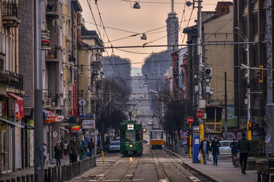 Dos tranvías, uno verde y uno amarillo, recorren las vías por el centro de una calle urbana llena de edificios y peatones.