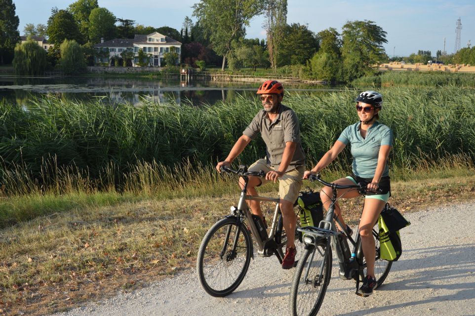 Un uomo e una donna con i caschi vanno in bicicletta su un sentiero sterrato lungo un lago con rigogliosi canneti verdi.