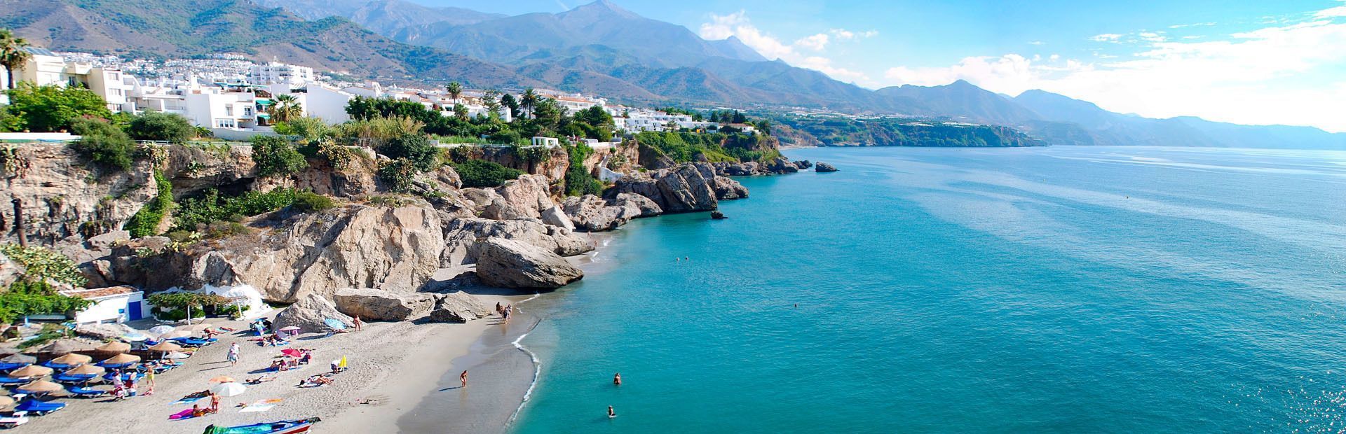 Edificios blancos sobre acantilados rocosos con vistas a una playa de arena y un mar turquesa, y montañas distantes bajo un cielo azul.
