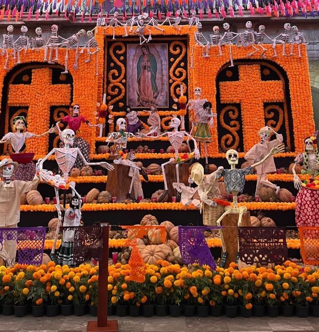 Un gran altar del Día de Muertos decorado con flores de cempasúchil naranjas, calabazas y numerosas calaveras calaca en varias poses.
