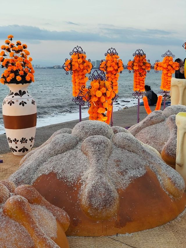 Una ofrenda del Día de Muertos en una playa con grandes esculturas de Pan de Muerto y cruces decoradas con cempasúchil naranja.