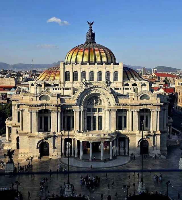 Un edificio neoclásico ornamentado con una cúpula de azulejos coloridos, visto desde arriba, con una plaza de la ciudad y montañas al fondo.