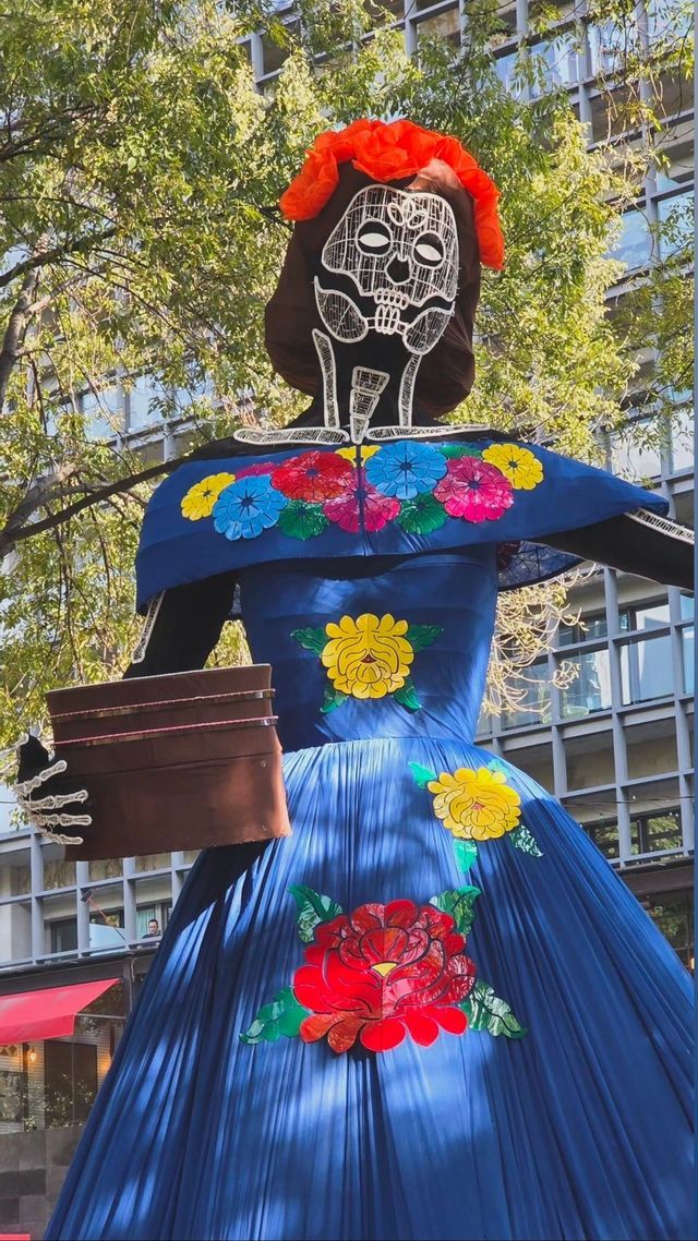 Una gran figura de Calavera Catrina con un rostro de calavera blanco y un vestido azul adornado con flores coloridas se encuentra al aire libre.