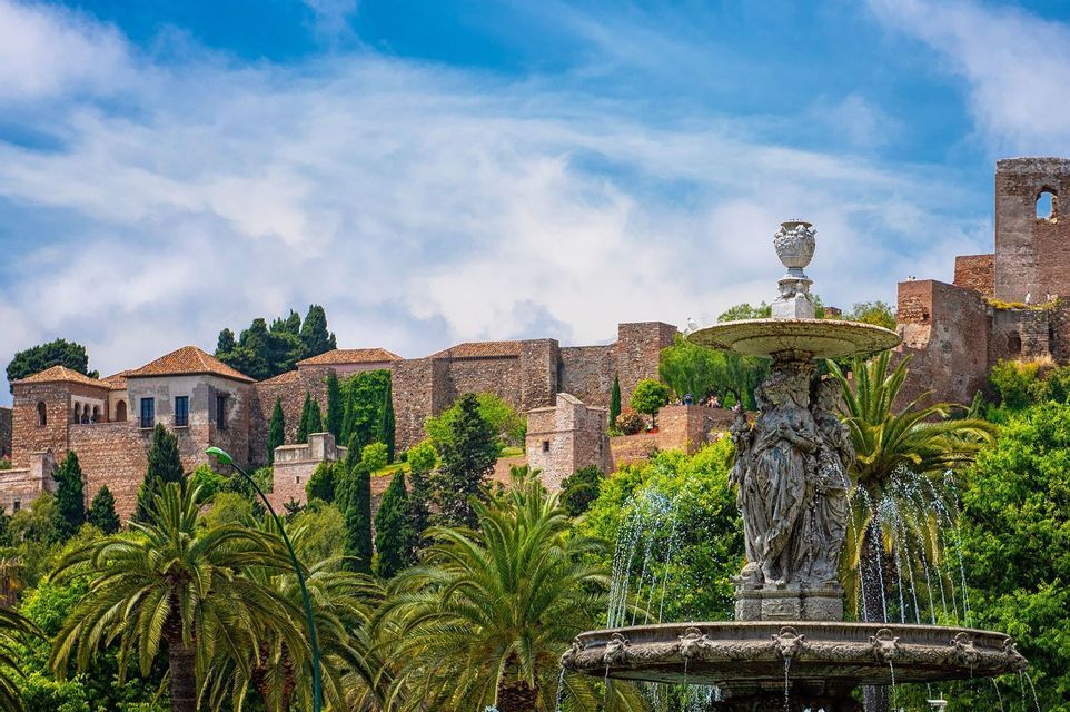 Una ornamentada fuente de piedra rocía agua en un exuberante parque, con una histórica fortaleza de piedra en una colina detrás bajo un cielo azul.