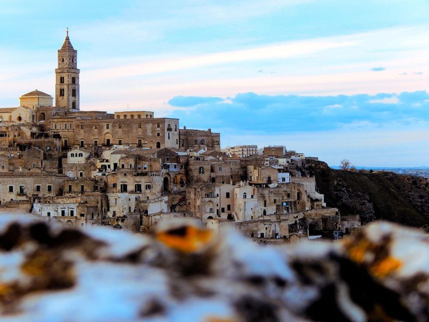 Un borgo in pietra con un campanile imponente, arroccato su una collina, visibile oltre un primo piano sfocato e sotto un cielo dai colori pastello.