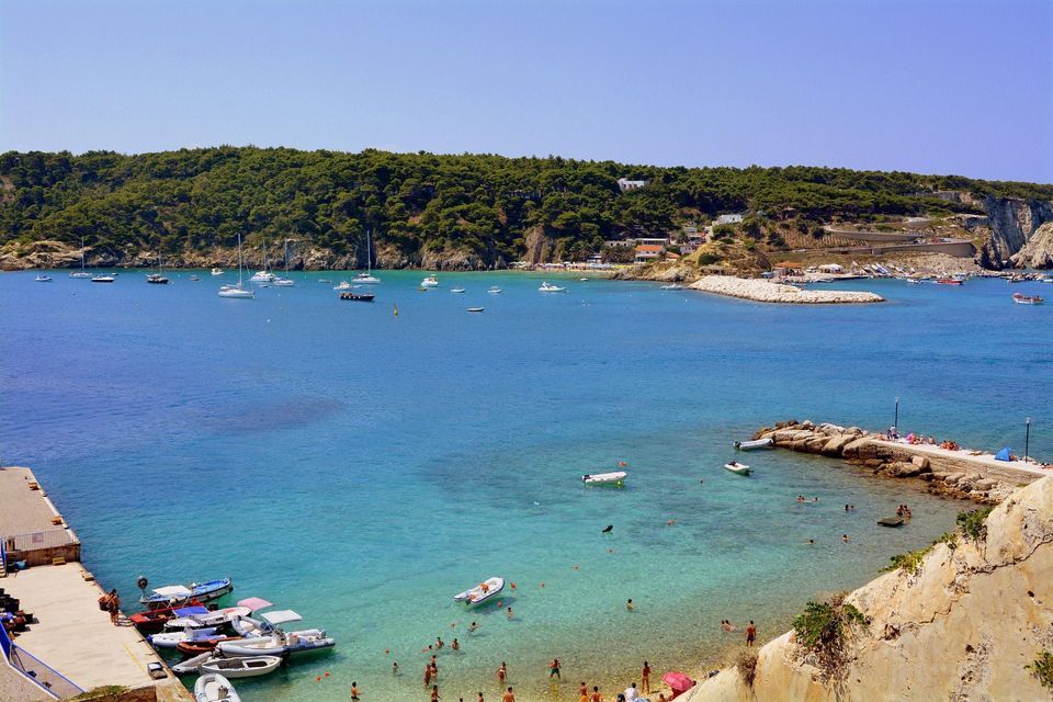 Una veduta dall'alto di una baia costiera dove le persone nuotano in acque turchesi, con barche ormeggiate a un molo e ancorate nel blu profondo del mare.