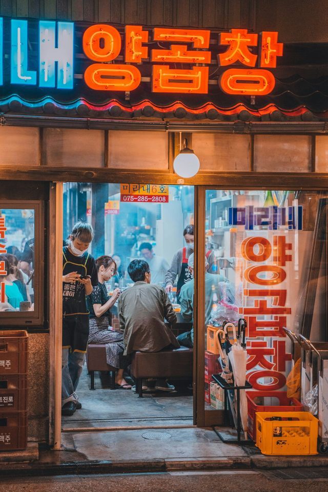 An open-front Korean restaurant bustling with customers at night, viewed from the street under glowing neon signs.