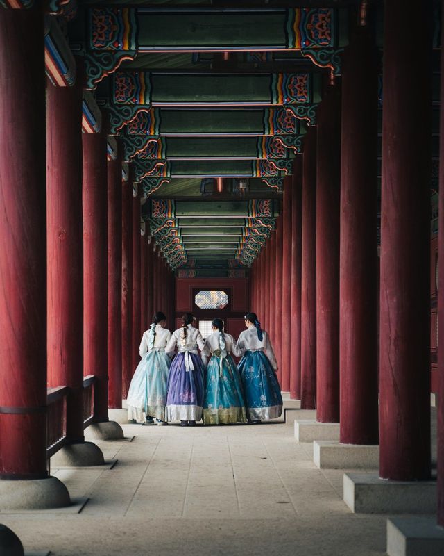 A WeRoad group trip of four women in colorful traditional hanbok dresses, seen from behind in a long corridor with red pillars.