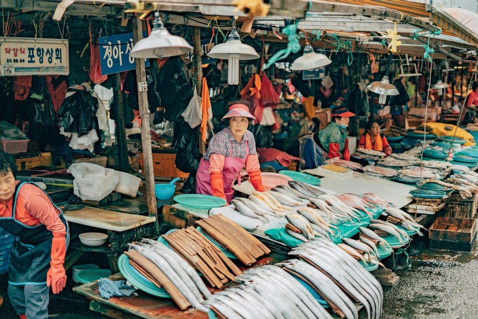 A female fish vendor in a pink apron and visor stands behind a stall filled with fresh fish at an outdoor market.