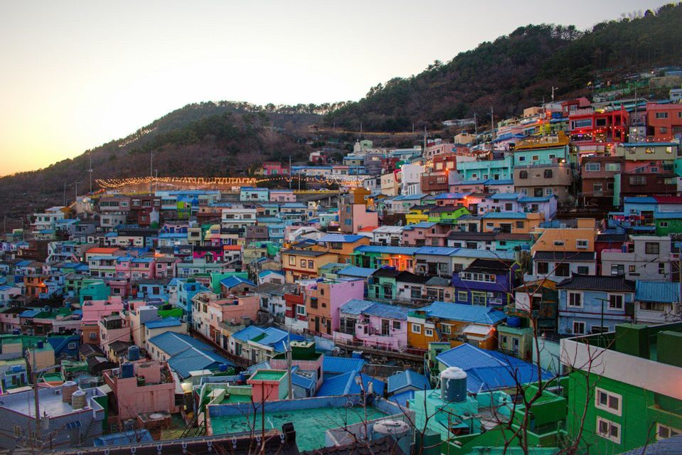 An overview of colorful, tightly packed houses built into a green hillside at sunset, with string lights illuminated.