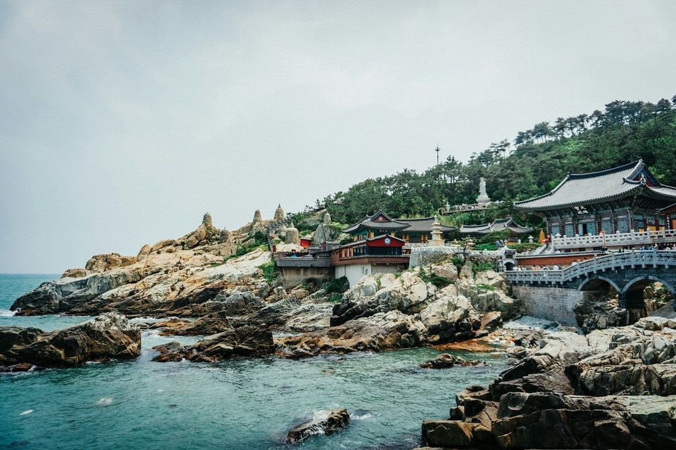A traditional temple complex built on a rocky coastline, with a stone bridge connecting buildings over the turquoise sea.