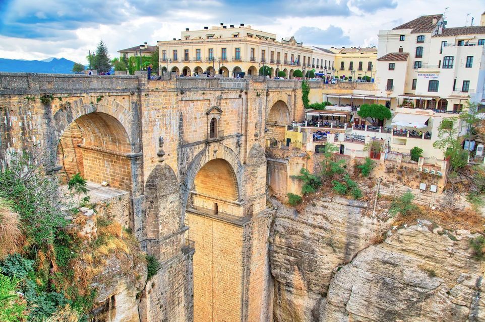 Un gran puente de piedra de múltiples arcos atraviesa un profundo desfiladero rocoso, conectando una ciudad histórica construida en los acantilados bajo un cielo nublado.